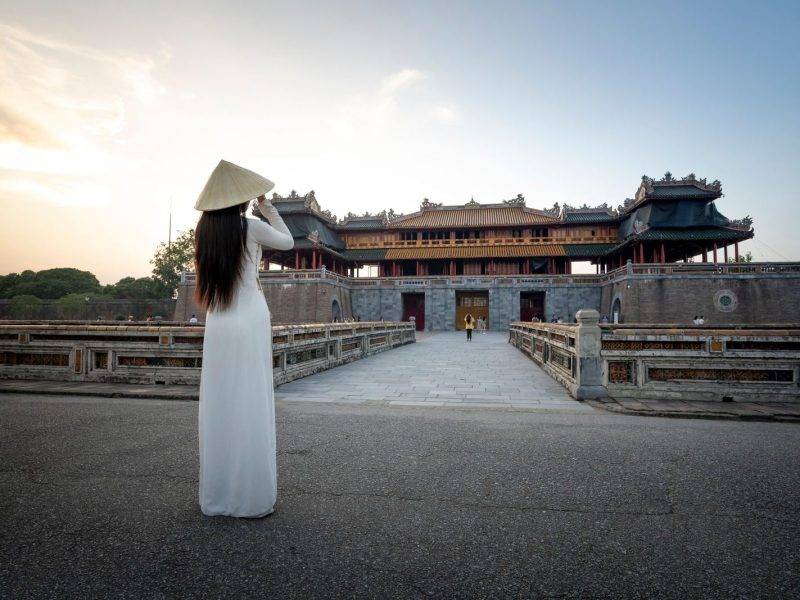 Back view full body of unrecognizable female in dress and hat standing against medieval meridian gate located in Hue city